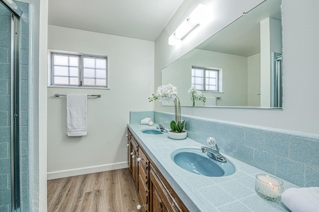 bathroom with wood finished floors, a shower with shower door, double vanity, and baseboards