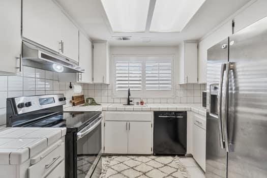 kitchen with appliances with stainless steel finishes, under cabinet range hood, a sink, tile countertops, and decorative backsplash