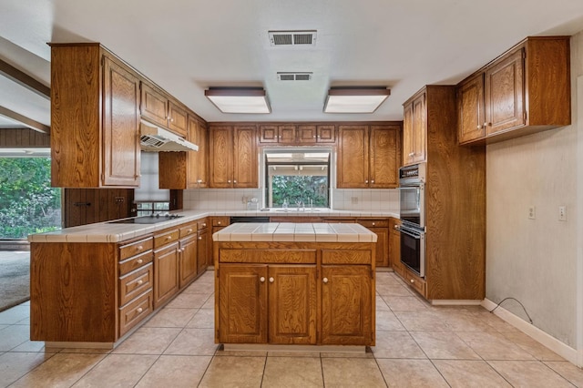 kitchen featuring tile counters, under cabinet range hood, tasteful backsplash, black electric cooktop, and brown cabinets