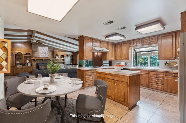 kitchen with tile countertops, under cabinet range hood, a large fireplace, ceiling fan, and a kitchen island