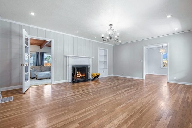 unfurnished living room with crown molding, light wood finished floors, a chandelier, built in shelves, and a warm lit fireplace