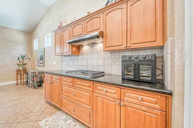 kitchen featuring under cabinet range hood, stainless steel gas cooktop, decorative backsplash, dark stone countertops, and light tile patterned floors