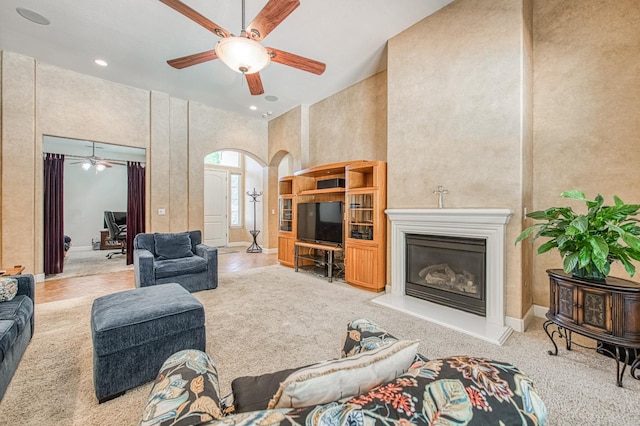 carpeted living room with ceiling fan, arched walkways, a high ceiling, a glass covered fireplace, and recessed lighting