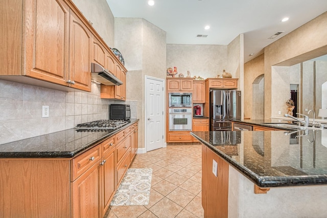 kitchen with stainless steel appliances, under cabinet range hood, backsplash, light tile patterned flooring, and dark stone countertops