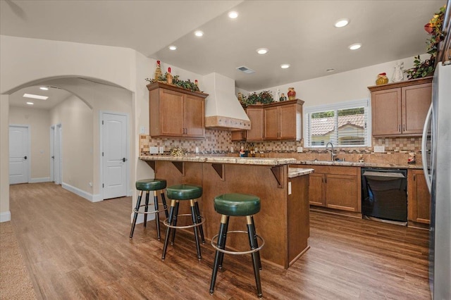 kitchen featuring dishwasher, custom exhaust hood, wood finished floors, brown cabinetry, and recessed lighting