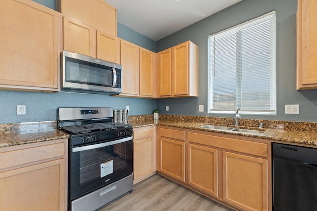 kitchen featuring appliances with stainless steel finishes, a sink, light brown cabinetry, light wood-style floors, and dark stone countertops