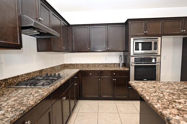 kitchen with appliances with stainless steel finishes, under cabinet range hood, light tile patterned floors, dark stone counters, and dark brown cabinetry