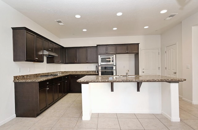 kitchen featuring stainless steel appliances, under cabinet range hood, dark stone countertops, a kitchen island with sink, and a sink