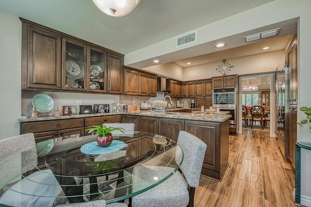 6772 N Wembley Dr, Fresno, CA 93711 - kitchen featuring wall chimney exhaust hood, light wood-type flooring, stainless steel double oven, light stone countertops, and glass insert cabinets