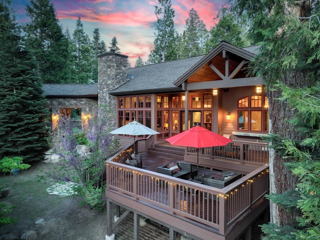 39516 Littleridge Rd, Shaver Lake, CA 93664 - back of house at dusk with a chimney, a shingled roof, a deck, stone siding, and outdoor dining area