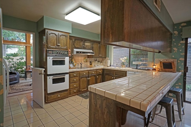 5513 N El Adobe Dr, Fresno, CA 93711 - kitchen with white appliances, under cabinet range hood, a peninsula, tile counters, and light tile patterned flooring