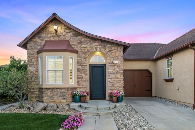 3613 Utah Ln, Clovis, CA 93619 - exterior entry at dusk featuring stone siding, an attached garage, concrete driveway, stucco siding, and a shingled roof