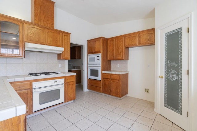 122 E Loop, Madera, CA 93637 - kitchen with white appliances, tile counters, under cabinet range hood, decorative backsplash, and glass insert cabinets