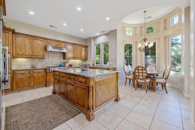 4638 W Lark Ct, Visalia, CA 93291 - kitchen with light stone countertops, brown cabinets, under cabinet range hood, a chandelier, and decorative backsplash