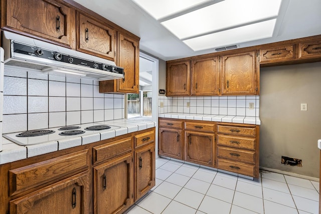 2076 W Calimyrna Ave Unit 102, Fresno, CA 93711 - kitchen with white electric cooktop, under cabinet range hood, tile counters, brown cabinets, and decorative backsplash
