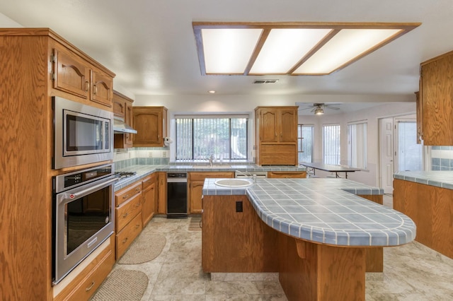 7664 N Kavanagh Ave, Fresno, CA 93711 - kitchen featuring stainless steel appliances, tile counters, under cabinet range hood, a kitchen island, and backsplash