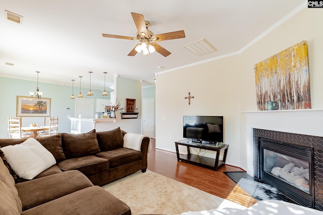 living room with crown molding, a tiled fireplace, wood-type flooring, and ceiling fan with notable chandelier