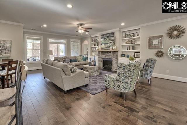 living room with built in features, ceiling fan, a stone fireplace, crown molding, and dark hardwood / wood-style floors