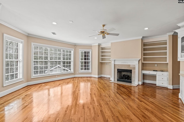 unfurnished living room with a tiled fireplace, light hardwood / wood-style floors, built in desk, ceiling fan, and ornamental molding