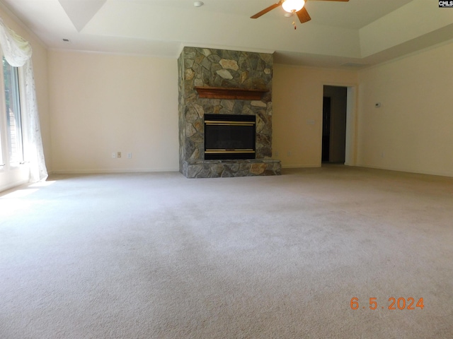 unfurnished living room with light carpet, a tray ceiling, a stone fireplace, and ceiling fan