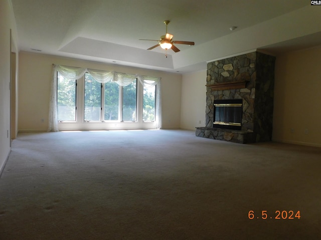 unfurnished living room with a tray ceiling, a stone fireplace, ceiling fan, and carpet flooring