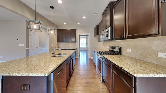 kitchen with appliances with stainless steel finishes, sink, hanging light fixtures, light stone counters, and a center island with sink