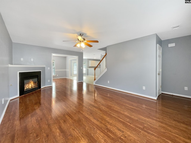 unfurnished living room featuring stairway, wood finished floors, baseboards, ceiling fan, and a glass covered fireplace