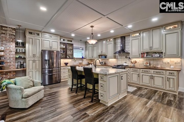 kitchen featuring hanging light fixtures, a kitchen island with sink, dark wood-type flooring, wall chimney range hood, and appliances with stainless steel finishes