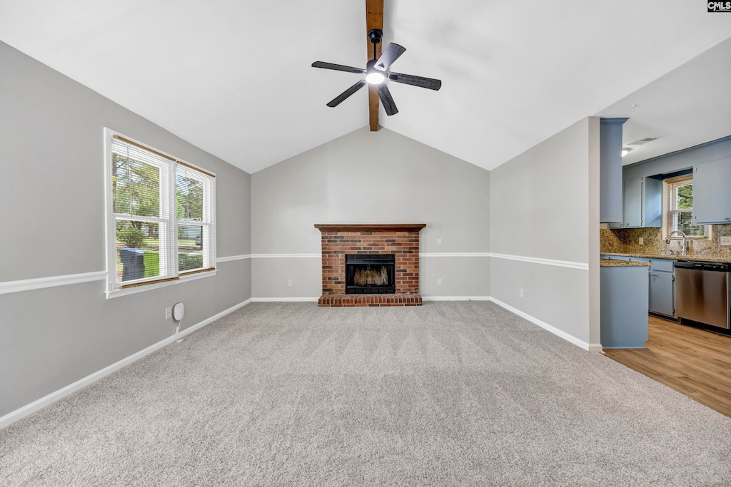 unfurnished living room featuring ceiling fan, vaulted ceiling with beams, sink, a brick fireplace, and light colored carpet