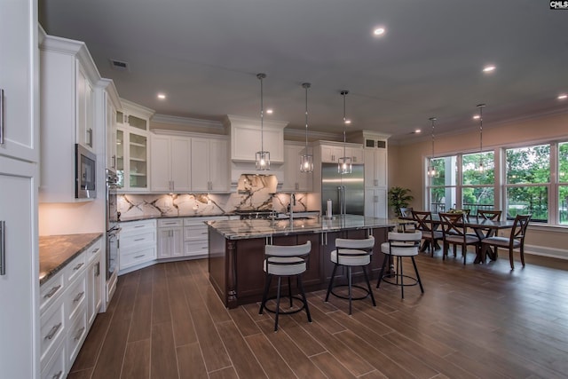 kitchen with a breakfast bar, dark wood-type flooring, a large island with sink, hanging light fixtures, and built in appliances