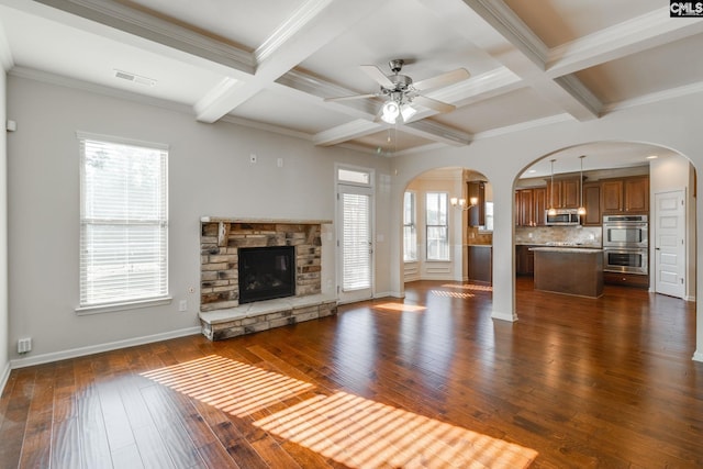 unfurnished living room featuring a stone fireplace, ceiling fan, beamed ceiling, ornamental molding, and dark hardwood / wood-style floors