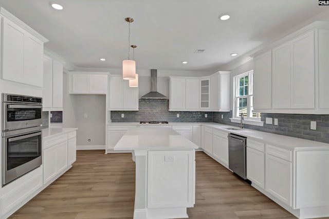 kitchen with a kitchen island, wall chimney range hood, stainless steel appliances, white cabinetry, and light hardwood / wood-style floors