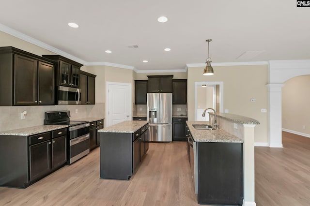 kitchen featuring stainless steel appliances, decorative light fixtures, a kitchen island with sink, ornate columns, and light wood-type flooring