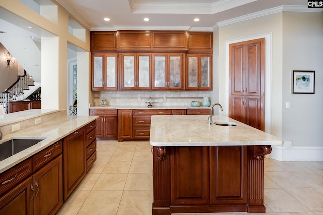 kitchen featuring sink, light stone counters, ornamental molding, backsplash, and an island with sink