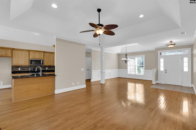 unfurnished living room with ceiling fan with notable chandelier, a raised ceiling, sink, light wood-type flooring, and ornate columns