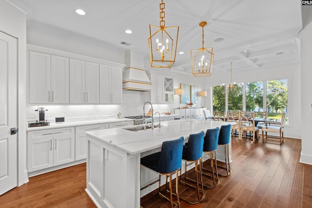 kitchen with premium range hood, a kitchen island with sink, white cabinets, decorative backsplash, and wood-type flooring