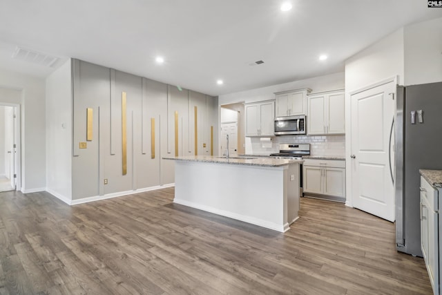 kitchen with light stone countertops, wood-type flooring, stainless steel appliances, and an island with sink