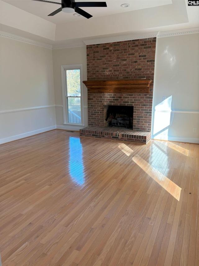 unfurnished living room featuring a raised ceiling, light wood-type flooring, a brick fireplace, and ceiling fan