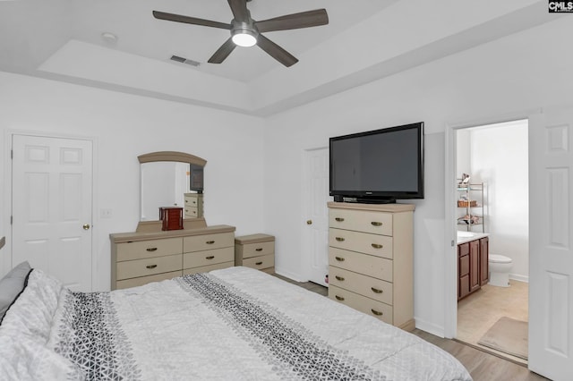 bedroom with a raised ceiling, light hardwood / wood-style floors, ensuite bath, and ceiling fan