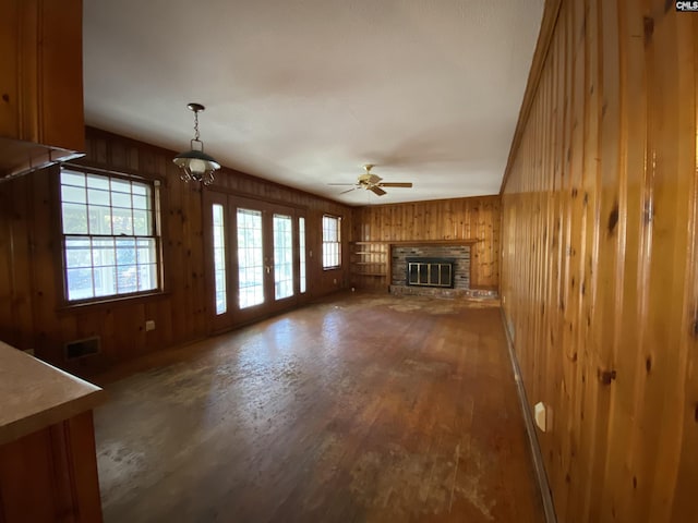 unfurnished living room featuring french doors, ceiling fan with notable chandelier, wooden walls, a brick fireplace, and hardwood / wood-style flooring