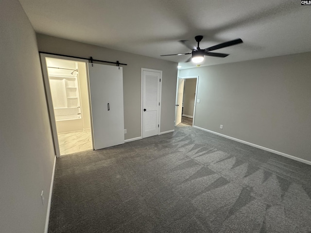 unfurnished bedroom with ensuite bath, ceiling fan, dark colored carpet, a barn door, and a closet