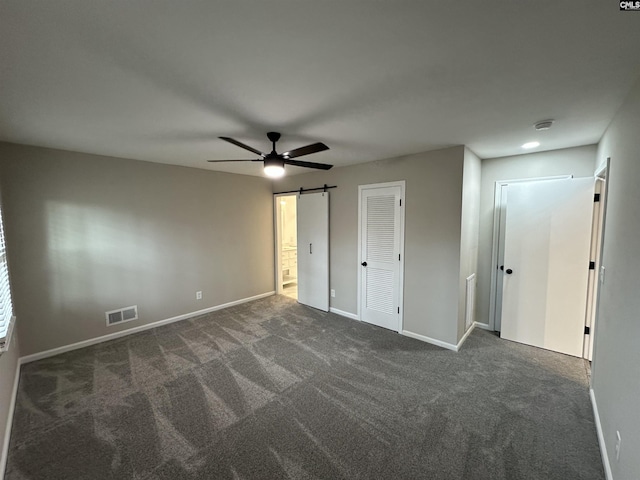 unfurnished bedroom featuring ceiling fan, a barn door, dark carpet, and two closets