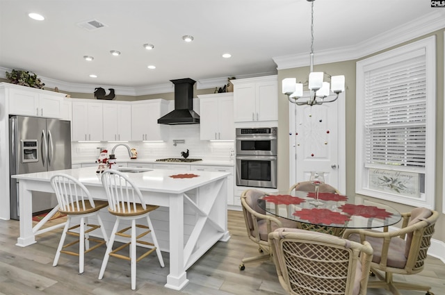 kitchen featuring wall chimney exhaust hood, stainless steel appliances, sink, a center island with sink, and an inviting chandelier