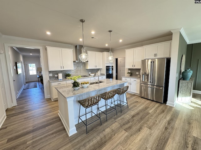 kitchen with light stone counters, stainless steel appliances, white cabinetry, an island with sink, and wall chimney exhaust hood