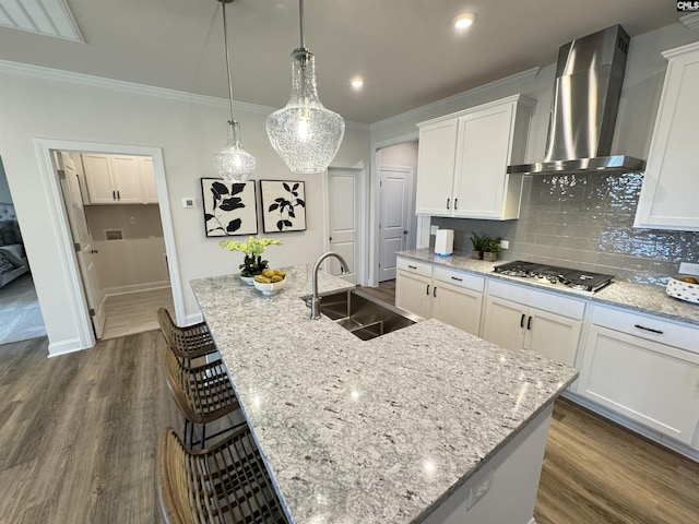 kitchen featuring a sink, white cabinets, wall chimney range hood, an island with sink, and pendant lighting