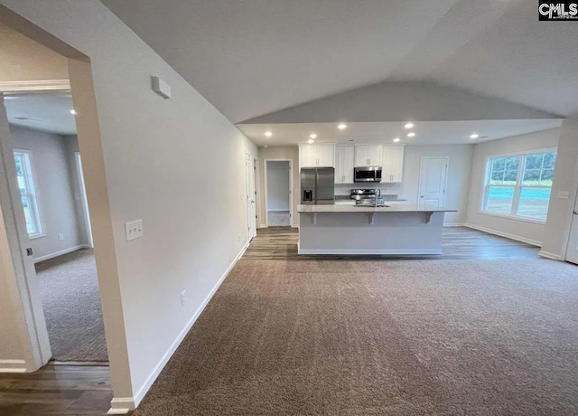 kitchen featuring dark colored carpet, stainless steel appliances, a kitchen island with sink, lofted ceiling, and white cabinets