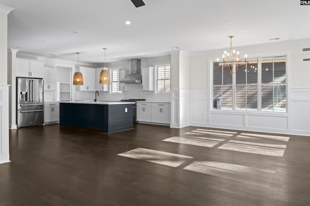 kitchen with wall chimney range hood, decorative light fixtures, a center island with sink, white cabinets, and appliances with stainless steel finishes