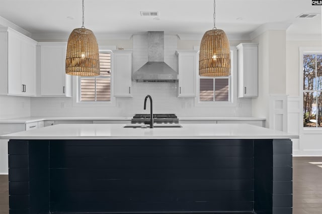 kitchen with hanging light fixtures, white cabinets, a kitchen island with sink, and wall chimney range hood