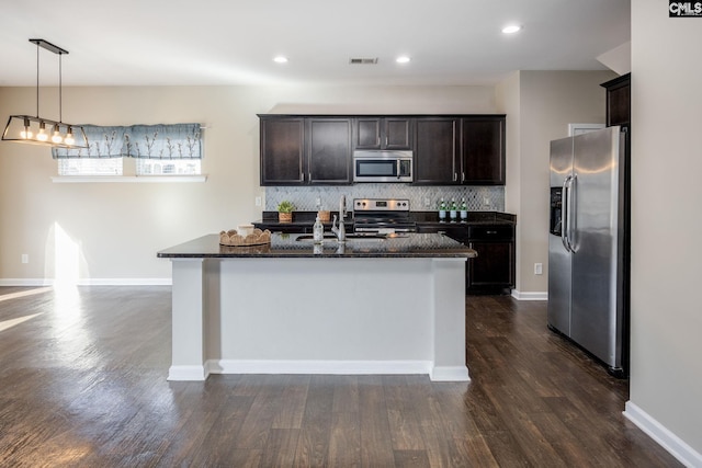 kitchen with decorative backsplash, appliances with stainless steel finishes, dark wood-type flooring, a center island with sink, and decorative light fixtures