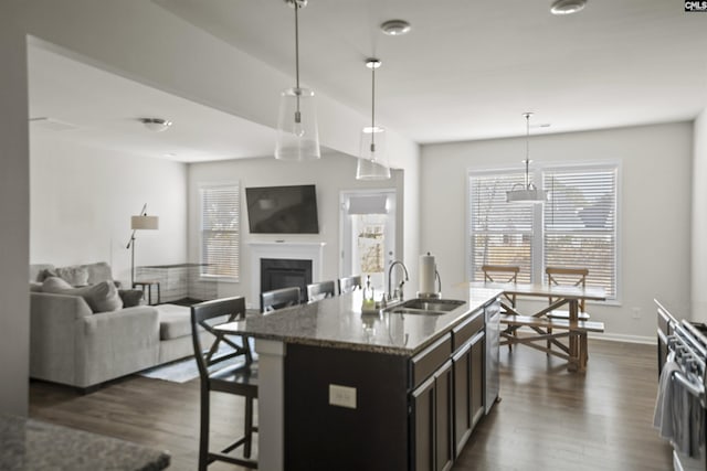 kitchen featuring a kitchen island with sink, hanging light fixtures, stainless steel dishwasher, light stone counters, and sink